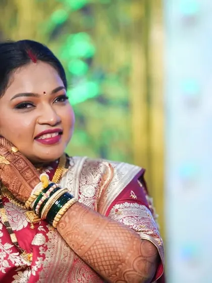 A candid moment capturing the beauty of a bride getting ready. Her makeup features a classic red bindi, defined eyes, and a happy smile. This look, with the intricate henna and green bangles, is the essence of a Maharashtrian bride.