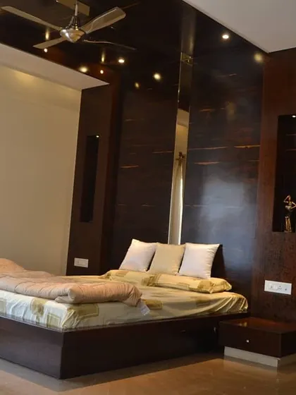 A sophisticated bedroom design featuring a floating bed and a full-height headboard wall panelled in a dark, high-gloss laminate. Integrated mirrors and a glossy false ceiling reflect light and add a sense of depth and luxury.