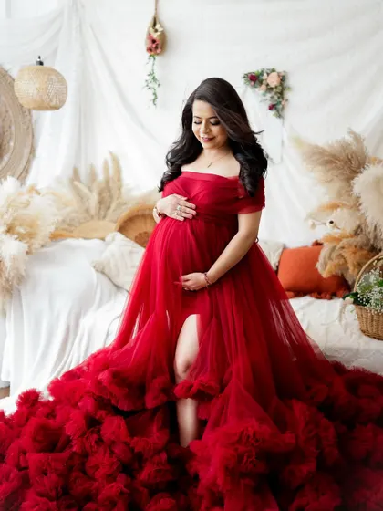 A stunning portrait of a mother-to-be in a vibrant red tulle gown. The boho-chic studio setup with pampas grass and soft lighting creates a warm and romantic atmosphere.