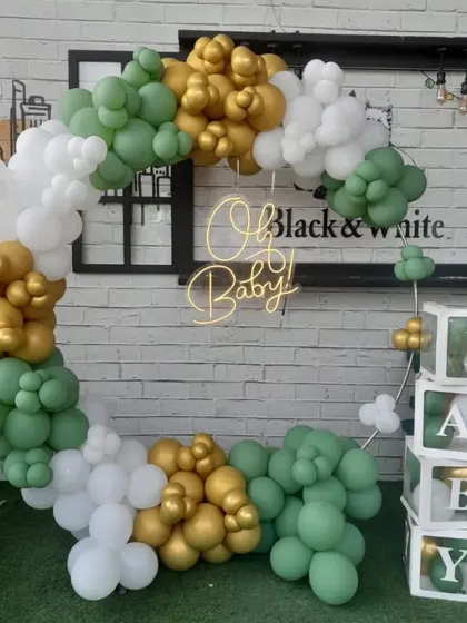 A modern and chic "Oh Baby" shower setup. The decoration features a stylish balloon arch in sage green, white, and gold, with a neon sign and "BABY" blocks.