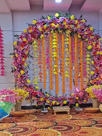 A colorful indoor Haldi or Mehendi setup with a circular floral arch and hanging tassels. The floor is covered with a traditional patterned rug, adding to the ethnic charm.