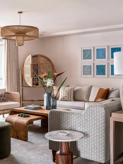 The living room reflects a relaxed, beachy vibe with an elegant, understated feel. The furniture includes a mix of slipcovered sofas, cane armchairs, and a unique burl veneer coffee table, all in a palette of soft neutrals and blues.