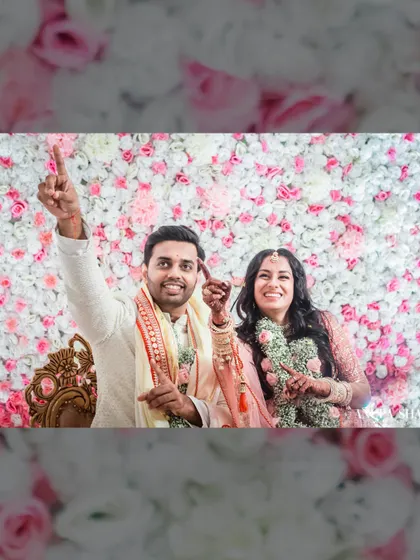 A fun, celebratory shot of the couple against a wall of flowers. They are pointing upwards, full of energy and joy after a wedding ritual.