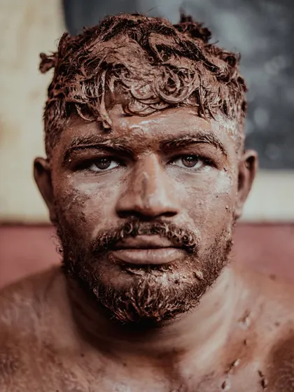 An intense close-up portrait of a wrestler, his face and hair caked with the reddish-brown soil of the taleem. His direct gaze conveys the focus and determination required for the sport.