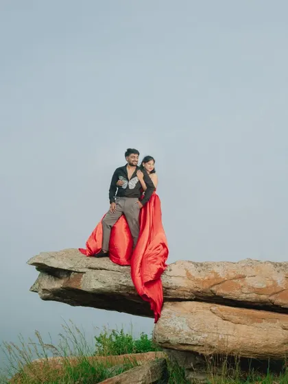 An epic and adventurous shot. The couple poses on a cliff edge, the red trail of her gown flowing dramatically below.