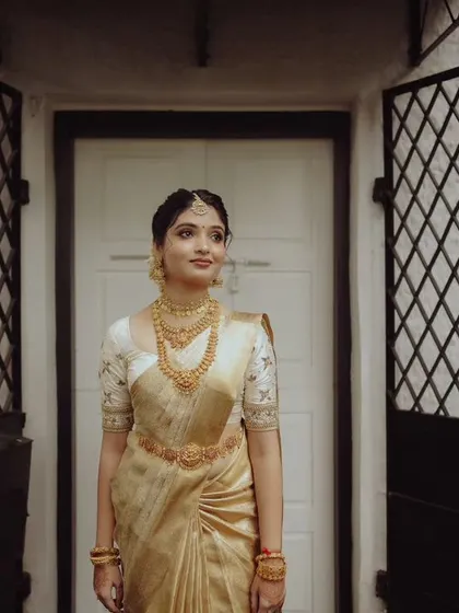 A classic bridal portrait of a woman in a cream and gold saree, standing elegantly in a doorway.