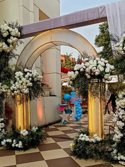 A grand entrance arch decorated with lush arrangements of white flowers and greenery. The golden pillars and soft lighting create a luxurious and welcoming feel for a sophisticated event.