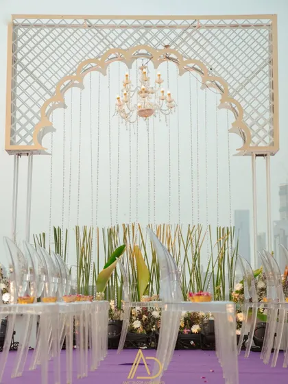 A modern seating arrangement with transparent chairs set against a backdrop of tall grasses and a beautiful white arch with a hanging chandelier.