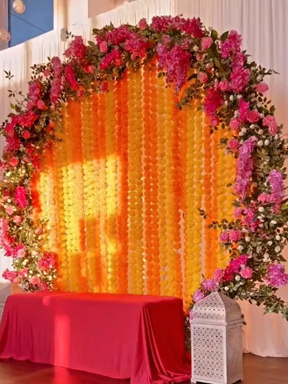 A circular backdrop for a Haldi or Mehendi ceremony, featuring a curtain of yellow and orange flower strings framed by a ring of lush pink flowers. This design creates a beautiful halo effect and is perfect for focusing attention on the main seating area.