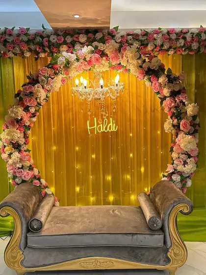 A glamorous Haldi backdrop featuring a circular arch of pink and white roses. A beautiful chandelier hangs in the center, and a 'Haldi' neon sign adds a modern twist.