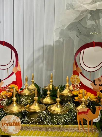 A detail shot of a Rakhi platter featuring elegant brass containers nestled in a bed of fresh flowers, accented with traditional dhol player cutouts for a celebratory feel.