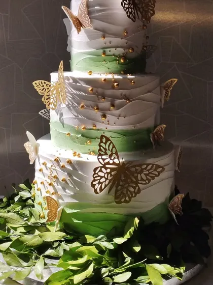 A stunning four-tiered cake with a green ombre effect and delicate gold butterflies. The base is surrounded by fresh green leaves, creating a beautiful natural aesthetic.