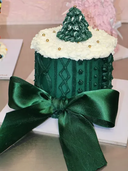 This cozy Christmas cake features a green sweater-knit texture and is topped with a snowy Christmas tree. The satin green bow makes it a perfect gift.