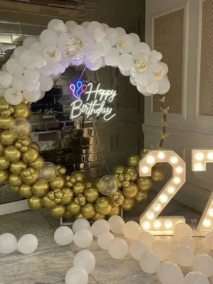 A chic 27th birthday setup featuring a semi-arch of gold and white balloons against a mirrored wall, paired with large marquee numbers.