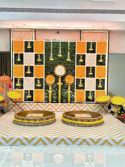 The main seating area for a Haldi, featuring a backdrop of checkered marigold panels and a chevron-patterned floor. The low traditional seating (bajots) completes the look.