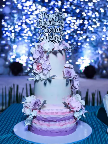 The 25th-anniversary cake in its element, set against a sparkling blue backdrop at the celebration. The silver "Happy 25th Anniversary" topper adds the final touch.