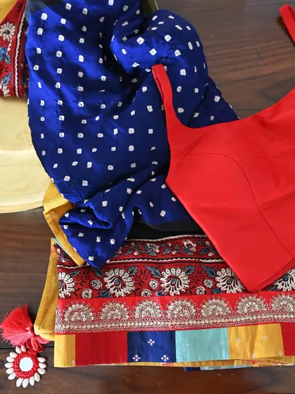A deconstructed view showing the components of a custom saree set. The deep blue Bandhni silk and its ornate Ajrakh border are paired with a simple, elegant red blouse to complete the ensemble.