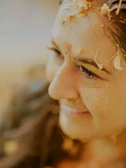 A close-up shot capturing the bride's serene smile during her haldi. Covered in turmeric paste and flower petals, she radiates a natural glow and happiness.