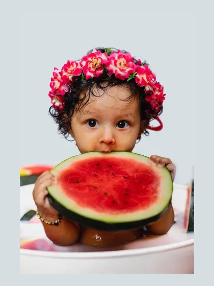 A baby girl holds a large slice of watermelon up to her mouth while sitting in a milk bath. This adorable photo perfectly encapsulates the fun of a themed baby shoot.