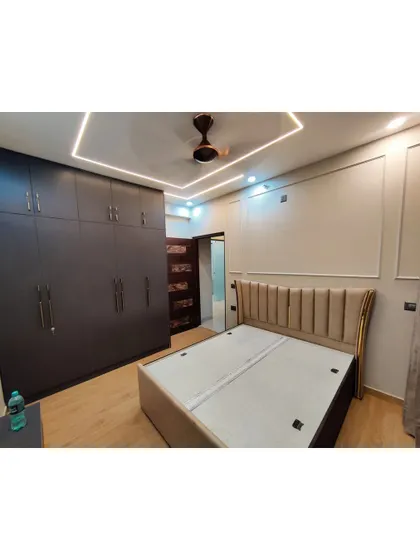 This bedroom showcases our custom wardrobe and bed design. The dark wood wardrobe provides ample storage, while the upholstered bed and modern false ceiling with cove lighting add a touch of luxury.