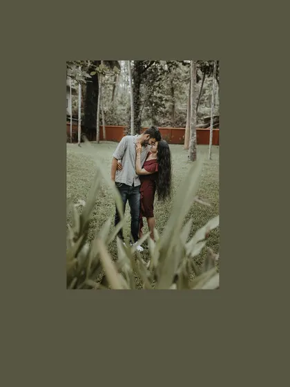 A full-length shot of a couple embracing in a park, framed by green leaves.