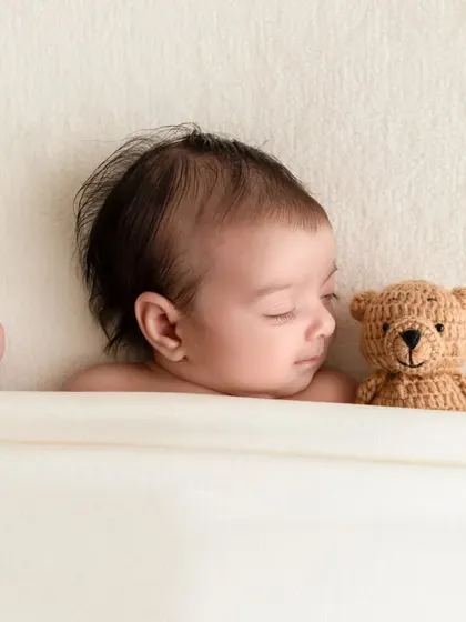 A sleeping newborn rests next to a small, knitted teddy bear, adding a sweet and classic touch to the portrait.