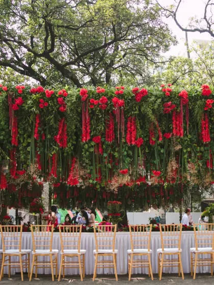 A long view of a dining table set under a spectacular hanging floral installation of red blooms and greenery, perfect for an elegant outdoor celebration.
