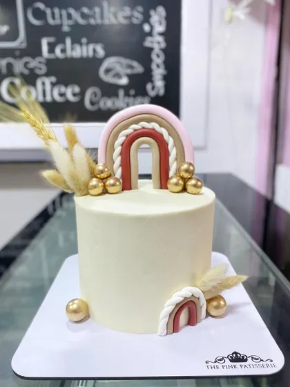 A trendy boho-themed cake with a neutral color palette. It features a fondant rainbow in earthy tones, dried wheat stalks, and gold sphere accents for a chic, rustic vibe.