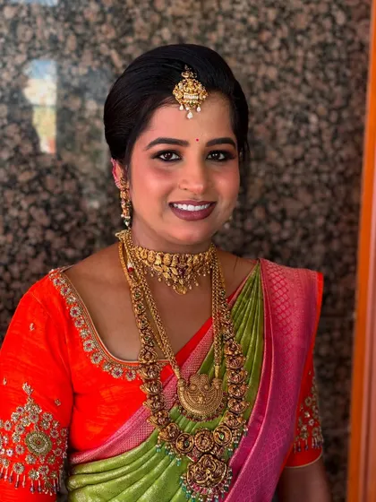 A smiling bride in a parrot green and pink saree, her hair in a neat updo that lets her jewellery shine.