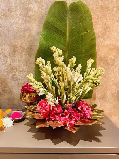 A stunning floral arrangement featuring white tuberoses and pink hydrangeas in a brass lotus bowl, set against a large banana leaf. This is a perfect example of our modern take on traditional floral design.