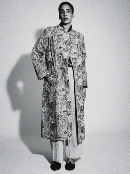 A full-length black and white portrait with strong, graphic lighting. This image has a minimalist, high-fashion feel, focusing on the silhouette of the long jacket.