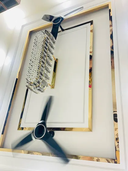 A creative shot of the ceiling, reflecting the chandelier and fans. It showcases the beautiful false ceiling design with its golden borders and cove lighting.