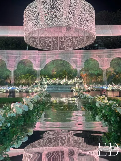 The reflection of the grand chandelier on the glossy black aisle creates a dramatic and mesmerizing effect. The path is flanked by abundant white floral arrangements, guiding the way to the main stage.
