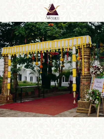 A traditional welcome arch with carved pillars and a canopy of yellow flowers. The red carpet and welcome board complete the classic and elegant entrance for a wedding.