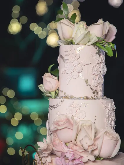 A dreamy wedding cake set against a backdrop of twinkling lights. The soft pink tiers are adorned with delicate lace piping and fresh roses, creating a truly romantic and elegant centerpiece.