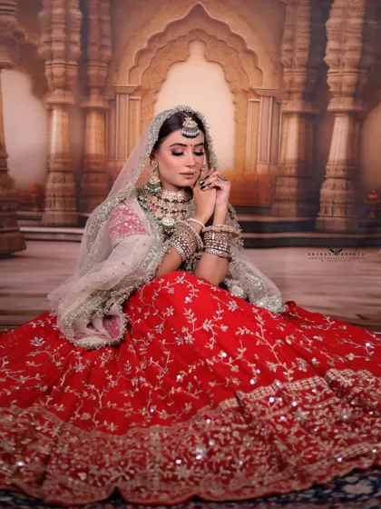 A full-length shot of the bride seated, with her voluminous red lehenga spread out before her. The palace-themed backdrop adds to the royal and grand feel of the photograph.
