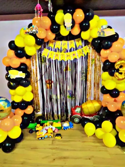 A construction-themed birthday party for a 3-year-old. The setup includes a black and yellow balloon arch, a silver fringe curtain, and various toy trucks and excavators as part of the decor.