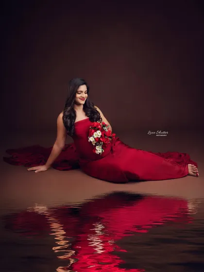 The reflection on the floor adds a dreamy, artistic quality to this portrait. The rich red of the gown contrasts beautifully with the dark, moody backdrop.