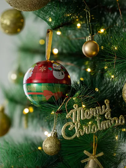 A close-up of a Christmas ornament, which is also a chocolate tin, hanging on a decorated tree. This highlights creative product usage for festive decor.