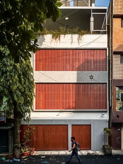 A minimalist urban residence in Basavanagudi, featuring a clean concrete frame with bold, red-orange vertical louvers that provide privacy and a strong visual identity.
