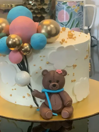 A cute teddy bear cake perfect for a baby shower or first birthday. The bear is holding a bunch of pink, blue, and gold balloons.