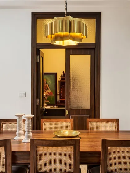 A classic dining setup with a modern twist. The brass chandelier adds a touch of mid-century style above the wood table and cane chairs, with a view into the home's puja room.