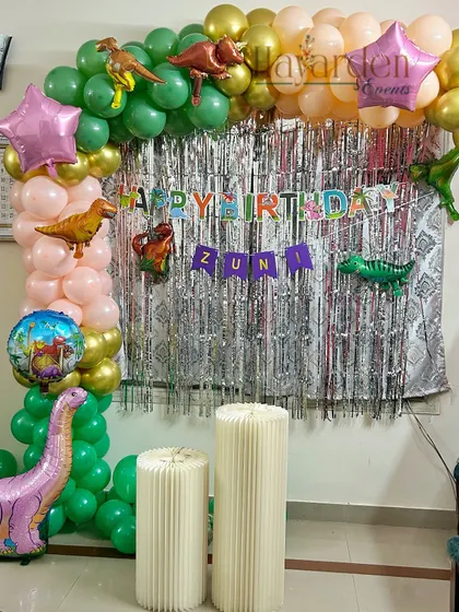 A dinosaur-themed birthday party with a green and peach balloon arch. Foil dinosaur balloons and a silver fringe curtain complete this prehistoric look.