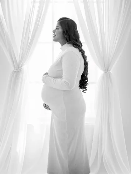 A serene black and white portrait taken against a window with sheer curtains. The soft, diffused light creates a gentle and timeless mood.