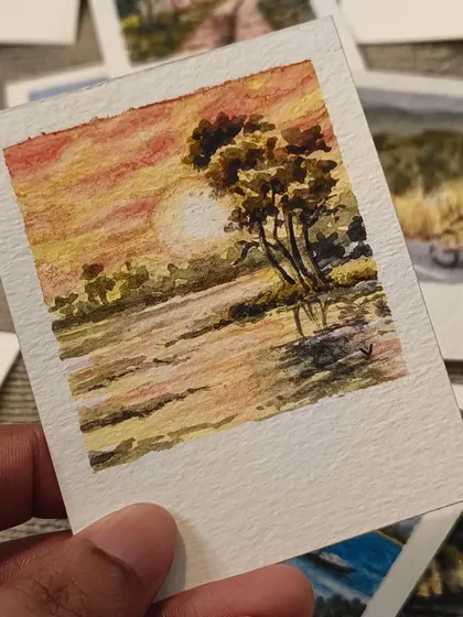 A warm, golden sunset over the water, painted in watercolor on a miniature Polaroid. The reflection of the sun and the silhouette of the tree create a peaceful, dreamy atmosphere.