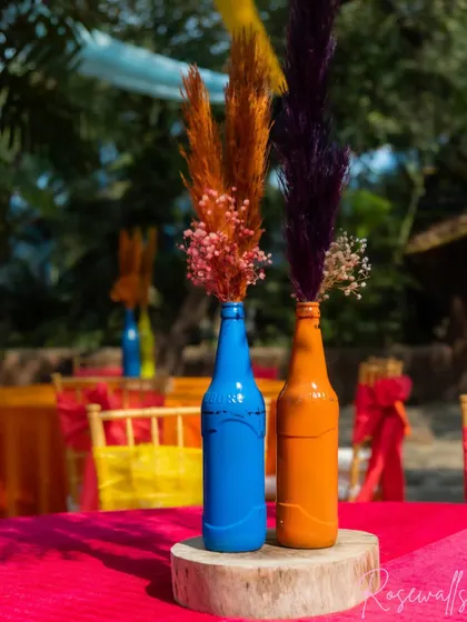 The centerpieces were simple yet effective, featuring colorful painted bottles with dried flowers, placed on a wooden slice for a rustic touch.