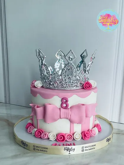 A cake fit for a princess! This lovely pink and white cake is adorned with a large silver crown, pink bows, and delicate roses for an 8th birthday celebration.