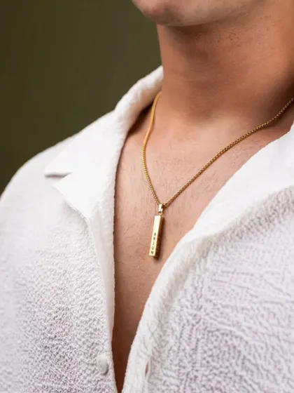 This lifestyle shot shows how a piece of jewelry complements personal style. The focus is on the gold pendant against a simple white shirt, demonstrating the product in a natural, wearable context for a men's accessory brand.