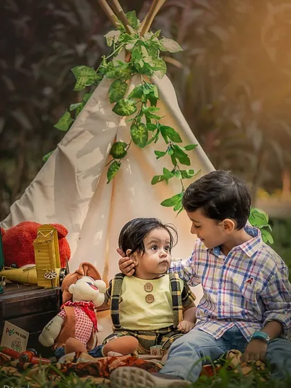 In a charming outdoor setup with a teepee, an older brother gently touches his baby brother's cheek. This is a beautifully composed shot that captures a tender moment of sibling care.