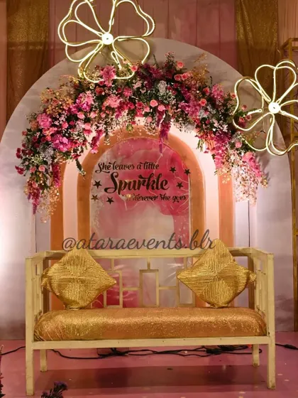 A beautiful and sparkling backdrop for an Aqeeqah celebration. The sign "She leaves a little sparkle wherever she goes" is set against a pink and gold arch, adorned with a lush floral arrangement.